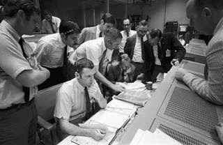 A group of flight controllers gathers around the console of Glenn S. Lunney (seated, nearest camera), Shift 4 flight director, in the Mission Operations Control Room (MOCR) of Mission Control Center (MCC), located in Building 30 at the Manned Spacecraft Center (MSC). Their attention is drawn to a weather map of the proposed landing site in the South Pacific Ocean. Among those looking on is Dr. Christopher C. Kraft, deputy director, MSC, standing in black suit, on right. When this photograph was taken, the Apollo 13 lunar landing mission had been canceled, and the problem-plagued Apollo 13 crew members were in trans-Earth trajectory attempting to bring their crippled spacecraft back home.<br /><br />Credits: NASA